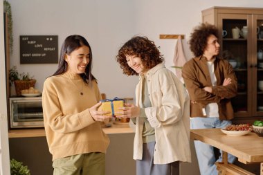 Gülümseyen kadın, hediye kutusuna sarılı bir şekilde en iyi arkadaşının önünde eğilerek hediye paketi veriyor. Mutfağın ortasında duruyorlar.