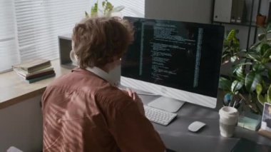 Genç erkek programcı kulaklık takıyor. Çalışma saatleri boyunca bilgisayarda kod yazıyor. Üzerinde kablosuz klavye, fare ve kahve fincanı var.