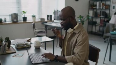 Afro-Amerikalı erkek programcı şirket ofisinde dizüstü bilgisayarla çalışırken müşteriyle telefon görüşmesi yapıyor.