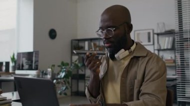 Afrikalı Amerikalı milenyum erkek programcısı ofiste dizüstü bilgisayar üzerinde çalışırken hoparlör modu kullanarak cep telefonuyla konuşuyor