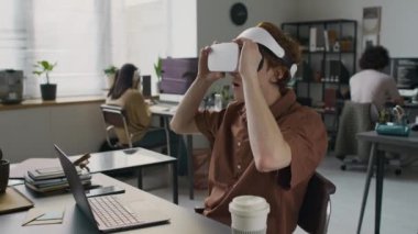 Kızıl saçlı genç erkek web mühendisi ofis masasındaki dizüstü bilgisayarda program yapmak için VR gözlük kullanıyor.