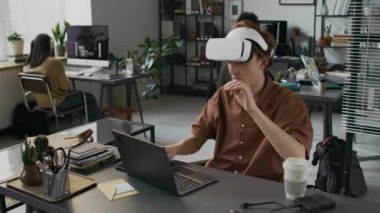 Gen Z erkek web geliştiricisi sanal gerçeklik kulaklığı kullanıyor ve bilgisayarında yazarken aynı zamanda bilişim şirketinde yeni bir sunucu için arayüz oluşturuyor.