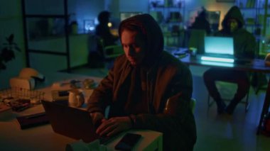 Siyah kıyafetli genç bir erkek siber hırsız karanlık ışıkta bilgisayara sızmak için kötü niyetli kod kullanıyor.