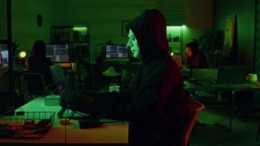 Siyah kapüşonlu beyaz maskeli profesyonel hacker gece geç saatlerde ofiste siber saldırı düzenlemek için dizüstü bilgisayar kullanıyor.