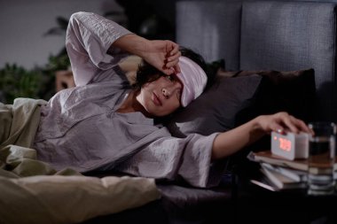 Yatağın yanındaki komodindeki alarmı durdurmak için ipek uyku maskesini çıkaran kadın.