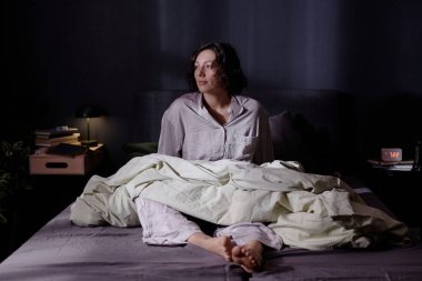 Rahat pijamalarıyla gülümseyen, bacaklarında zeytin battaniyesiyle yatakta oturan ve ışık saçan kadın.