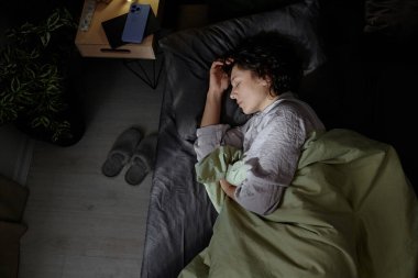 Yüksek açılı bir çekimde kadın uyurken ve yumuşak bir battaniyeye sarılırken ilk güneş yatağını aydınlatırken.