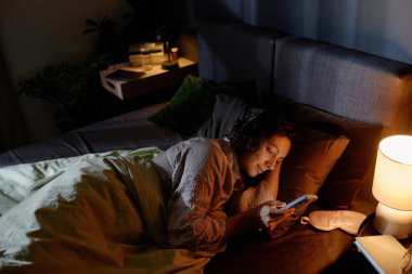Yüksek açılı çekimde, esmer kadın yatakta yatarken ve yatakta uykusuzluk çekerken akıllı telefon kullanırken görülüyor.