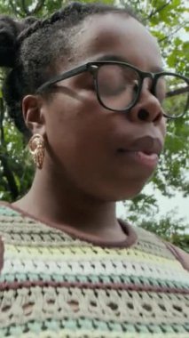 Siyah kadının kolsuz bluz giydiği, gözlüğünü çıkardığı, yaz günü parkta oturduğu el kamerası görüntüsü.