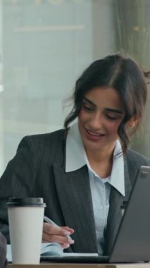Gri ceketli, beyaz gömlekli, yüzü tanınmayan bir arkadaşla konuşan ve laptopunu kafede gösteren gülümseyen bir kadın.