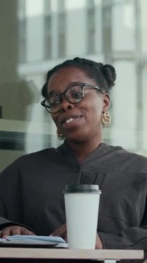 Siyahi bir kadının koyu renk kıyafet giydiği, konuşurken el kol hareketi yaptığı, kahve molası sırasında kafede oturduğu dikey bir fotoğraf.