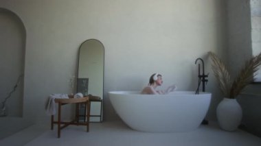Kulaklıklı genç bir kadın otelin minimalist banyosundaki beyaz oval küvette sabahtan zevk alıyor.