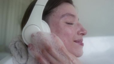 Kafasının altında havluyla küvette yatarken, beyaz kulaklıklarla müzik dinleyen pozitif nesil Z kızına yakından bakın.