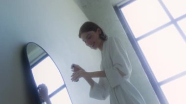 Geniş banyoda aynanın önünde dikilirken beyaz bornoz giymiş, yanağına damla serumu süren neşeli genç bir kadının düşük açılı görüntüsü.