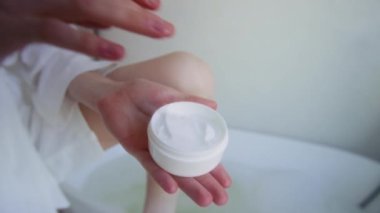 Banyoda cilt bakımı yaparken beyaz kavanozdan bir damla krem alan tanınmayan genç bir kadının yakın plan görüntüsü.