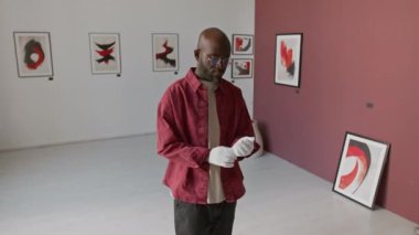 Özgüvenli Afrikalı Amerikalı bir adamın portresi. Eldivenlerini ayarlıyor ve kollarını kavuşturuyor. Minimalist sanat galerisinin ortasında soyut resimlerle dikilirken kameraya bakıyor.