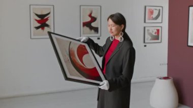 Sıradan giyinmiş Asyalı kadın ve beyaz eldivenler dikkatle tutuyordu ve çerçevelenmiş soyut sanat eserlerini inceliyordu, minimalist sanat galerisinde duruyordu.