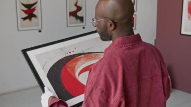 Beyaz eldiven giyen kel Afrikalı Amerikalı adam çerçeveli sanat eserleri taşıyor. Minimalist sanat galerisindeki duvarlarda diğer soyut resimlerle çevrili.