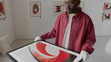 Afro-Amerikan bir adamın günlük kıyafetleri ve beyaz eldivenleriyle iki eliyle çerçeveli soyut sanat eserlerini tutarken, minimalist sanat galerisinde dikilirken.