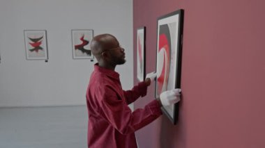 Beyaz eldivenler giyen Afrikalı Amerikalı adam, minimalist sanat galerisindeki duvara özenle soyut resim asıyor.