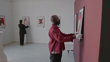 Afrikalı Amerikalı erkek ve Asyalı kadın, duvarlara soyut resimler dağıtarak minimalist sanat galerisini sergi için hazırlıyorlar.
