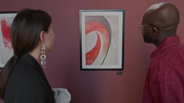 Asyalı kadın ve siyah adam duvarda soyut bir tabloyla karşı karşıyalar ve bunu tartışıyorlar, minimalist sanat galerisinde birlikte duruyorlar.