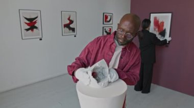 Afrikalı Amerikalı bir adam pedesnal 'a özenle soyut bir kurgu yerleştirirken, kadın meslektaşı da minimalist sanat galerisindeki duvara resim çiziyor.