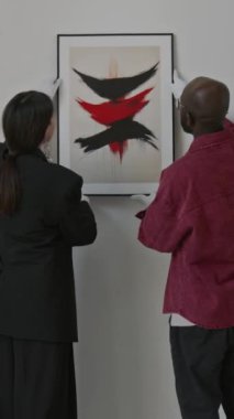 Kadın sanat küratörü ve asistanının modern sanat galerisindeki sergi için soyut resimleri duvara asmalarının dikey görüntüsü.
