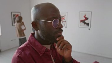 Gözlüklü, sakallı Afrikalı Amerikalı bir adamın, galeride soyut resimlerle sergilenen modern sanat eserlerini incelerken yakın plan çekimleri.