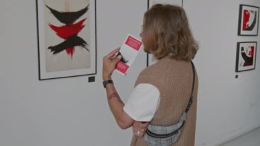 Uzun saçlı, örgü kazaklı bir adam farklı modern soyut tabloları inceliyor, galeride sanat sergisi broşürü tutuyor.