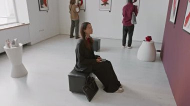 Asyalı bir kadının tablolara baktığı yüksek açı, iki adam ayakta dikilirken ve galeride diğer sanat eserlerini izlerken...
