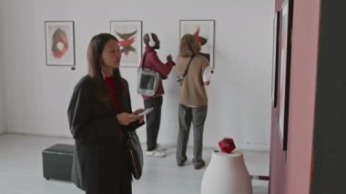 Asyalı kadın akıllı telefonuyla modern soyut resmin fotoğrafını çekerken, iki erkek minimalist galeride diğer sanat eserlerini tartışıyor.