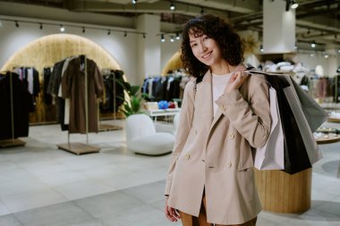 Esmer, kıvırcık saçlı, genç, moda düşkünü, çantasının arkasında kağıt torbalarla butikte poz verirken samimi bir şekilde gülümseyen kadın portresi.