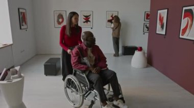 Asyalı kadın tekerlekli sandalyedeki zenciye yardım ediyor, galerideki sanat sergisinde soyut resimleri görmesi için onu yaklaştırıyor.