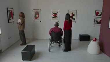 Tekerlekli sandalyedeki siyah adam ve diğer iki kişi minimalist galerideki sanat sergisinde soyut resimlere bakıyor.