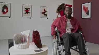 Diğer insanlar minimalist sanat galerisindeki duvarlardaki tablolara hayranlık duyarken, tekerlekli sandalyedeki Afrikalı Amerikalı adam, heykelin soyut yapısını incelemek için yaklaşıyor.