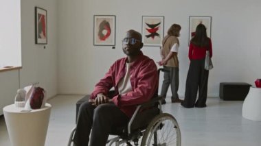 Tekerlekli sandalyede kameraya bakan Afrikalı Amerikalı bir adamın portresi. Minimalist galerideki diğer insanlar arasında soyut sanat sergisine katılıyor.