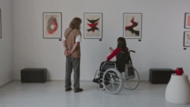 Tekerlekli sandalyedeki kadının ve uzun saçlı adamın galerideki sergide soyut sanatı tartışırken çekilmiş el kamerası görüntüsü.