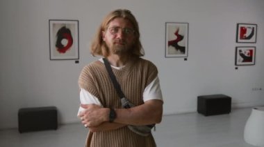 Minimalist galeride kollarını kavuşturmuş kameraya bakan uzun saçlı beyaz adamın portresi.