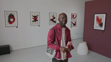 Minimalist galeride soyut resimlerle sergilenirken, elinde broşür tutan ve gülümseyen Afrikalı Amerikalı bir adamın portresi.