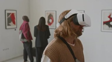 Uzun saçlı, VR kulaklık takan, dijital sanata bakan bir adam, diğer insanlar ise sanat galerisindeki duvarlardaki soyut kırmızı ve siyah tablolara hayran.