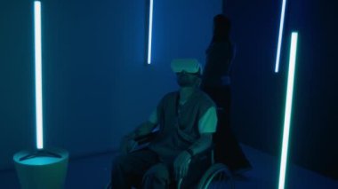 Tekerlekli sandalyede VR kulaklık takan bir adam loş ışıklı, dikey mavi ışıklı fütüristik bir odada otururken Asyalı bir kadın enstalasyonlara hayran.