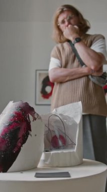 Modern galerideki sanat sergisinde uzun saçlı beyaz adamın düşünceli bir şekilde soyut heykele bakışının dikey görüntüsü.