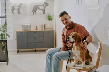 Çağdaş bir ofis odasında, duvarları ve bitkileri olan bir köpeğin yanında oturmuş gülümseyen genç adam rahat bir atmosfer yaratıyor.