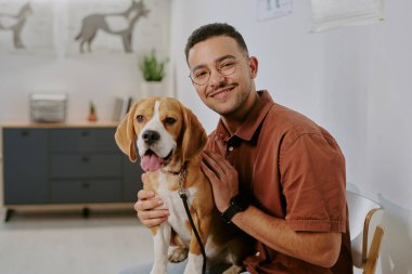 Gülümseyen bir adamın portresi, arka planda tıbbi diyagramlarla bir veteriner ofisinde oturan ve bir köpeği tutan bir adam.