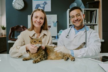 Gülümseyen veteriner ve kedi sahibi rahat bir kediyi muayene masasında tutarken tıbbi malzemeler ve duvar resimleri arka planda rahat bir klinik ortamını resmediyor.