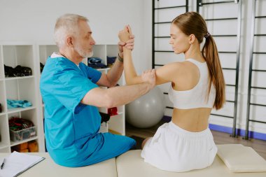 Back view shot of female patient sitting still while mature doctor fixing her hand to test flexibility of elbow joint clipart