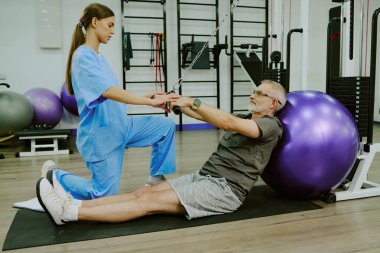 Yaralı yaşlı bir adamla egzersiz yapan genç bir kadın rehabilitasyon uzmanı, mindere oturup topa yaslanıyor.