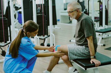 Rehabilitasyon merkezinde spor salonunda bankta oturan yaşlı bir hastanın dizini bantlayan genç bir koçun yüksek açılı çekimi.