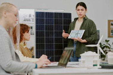Sınıfta laptoplarda çalışan meslektaşlarıyla yenilenebilir enerji modeli sunan genç bir kadın. Sürdürülebilir enerji uygulamalarına odaklan